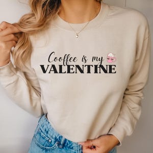 May include: Beige sweatshirt with the text "Coffee is my Valentine" in black script and block letters. A pink mug with whipped cream and a heart design is to the right of the text. The sweatshirt has long sleeves and a crew neck.