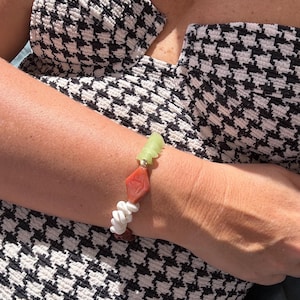 May include: A beaded bracelet featuring a mix of light green translucent glass beads, a central diamond-shaped orange bead, and white spiral shell beads. The bracelet is worn on a wrist against a black and white houndstooth patterned garment.