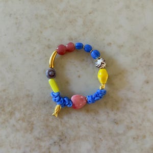 May include: A vibrant beaded bracelet with a gold fish charm. The bracelet incorporates red, blue, yellow, and white beads, alongside a pink heart-shaped bead. The bracelet is strung on a blue cord.