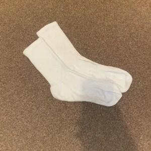 Puede incluir: Un par de calcetines blancos, hechos de un material suave y texturizado. Los calcetines están doblados y reposan sobre una alfombra marrón. Los calcetines tienen un diseño acanalado en la parte superior y un acabado liso en el pie.