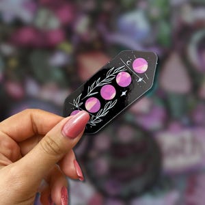 May include: A black sticker featuring a celestial design with three pink moon phases, white stars, and botanical accents. The sticker is held in a hand with pink nail polish, set against a blurred background.