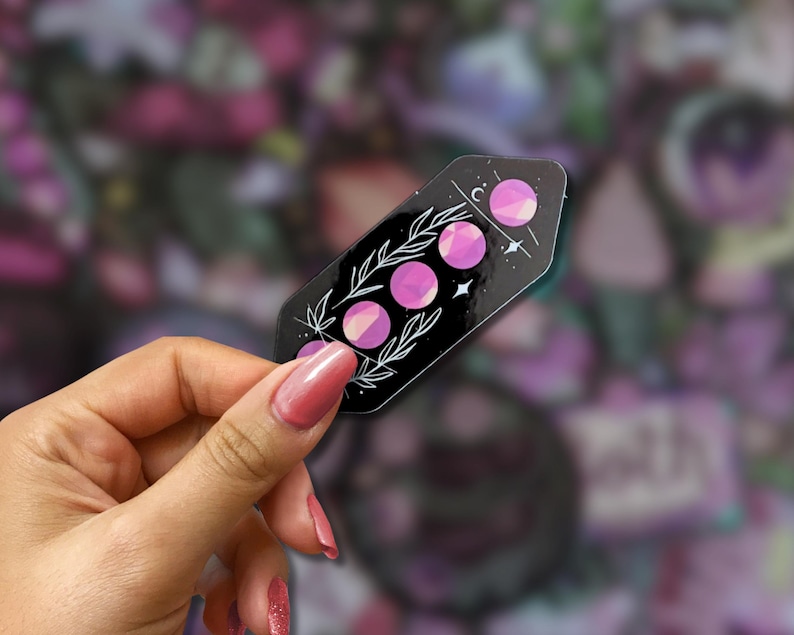 May include: A black sticker featuring a celestial design with three pink moon phases, white stars, and botanical accents. The sticker is held in a hand with pink nail polish, set against a blurred background.