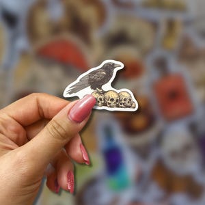 May include: A sticker featuring a black raven perched atop three skulls. The raven is detailed with textured shading, and the skulls are off-white. The sticker is held against a blurred background of other stickers.