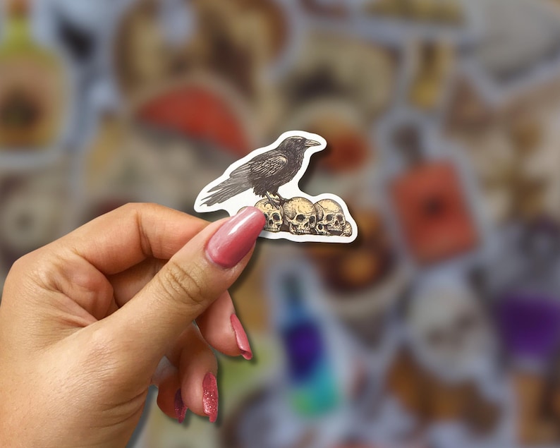 May include: A sticker featuring a black raven perched atop three skulls. The raven is detailed with textured shading, and the skulls are off-white. The sticker is held against a blurred background of other stickers.