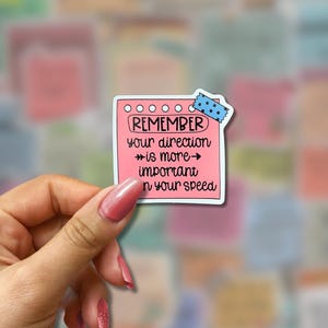 May include: A pink sticker in the shape of a notepad with the words "REMEMBER your direction is more important than your speed" in black text. The sticker is held by a hand with pink nail polish, and a blue polka dot tape is at the top.