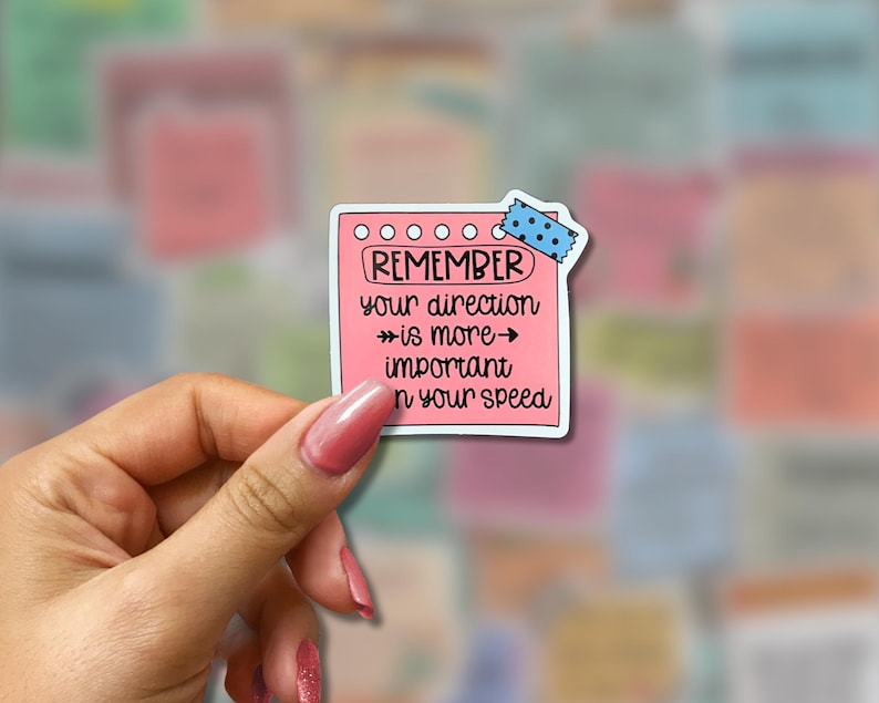 May include: A pink sticker in the shape of a notepad with the words "REMEMBER your direction is more important than your speed" in black text. The sticker is held by a hand with pink nail polish, and a blue polka dot tape is at the top.