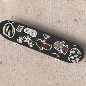 May include: Black skateboard with various white and black stickers. Stickers include stars, a heart with faces, and the word "ROCK". The skateboard has white wheels and is on a textured surface.