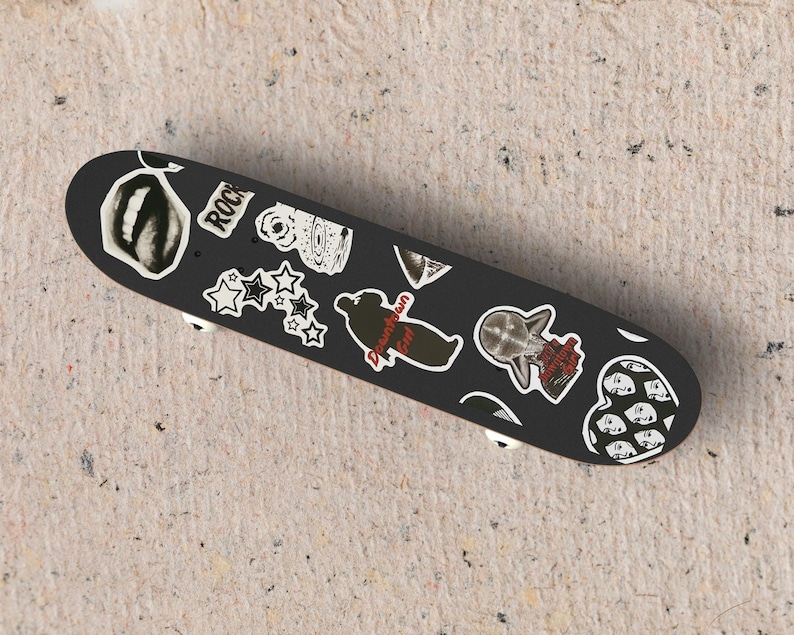 May include: Black skateboard with various white and black stickers. Stickers include stars, a heart with faces, and the word "ROCK". The skateboard has white wheels and is on a textured surface.