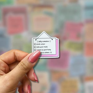 May include: A white sticker with a pink border and a small triangle at the top, resembling a hanging sign. The sticker lists daily reminders: drink water, take your meds, listen to your body, and be kind to others.