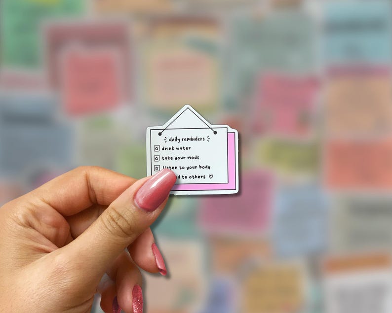 May include: A white sticker with a pink border and a small triangle at the top, resembling a hanging sign. The sticker lists daily reminders: drink water, take your meds, listen to your body, and be kind to others.