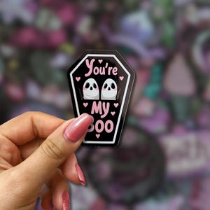 May include: A black coffin-shaped sticker with the text "You're My Boo" in pink. The sticker features two white ghost illustrations with black eyes and pink hearts. The sticker is held in a hand.