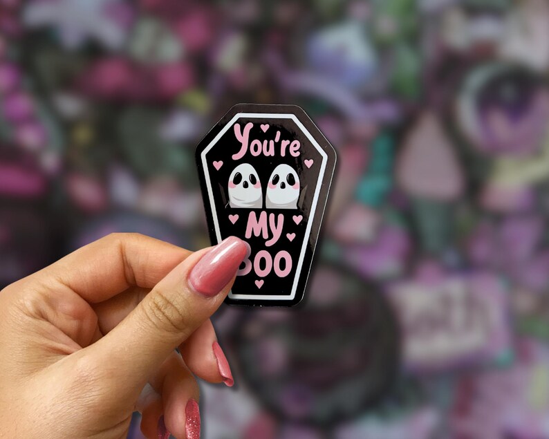 May include: A black coffin-shaped sticker with the text "You're My Boo" in pink. The sticker features two white ghost illustrations with black eyes and pink hearts. The sticker is held in a hand.