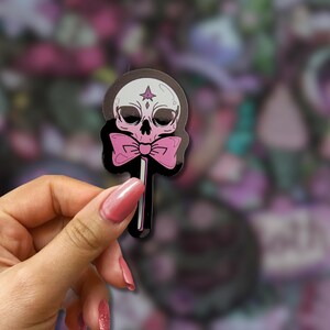 May include: A sticker featuring a skull-shaped lollipop with a pink bow tie. The skull is white with black eye sockets and a star on its forehead. The lollipop stick is black, and the background is a blur of purple and pink.