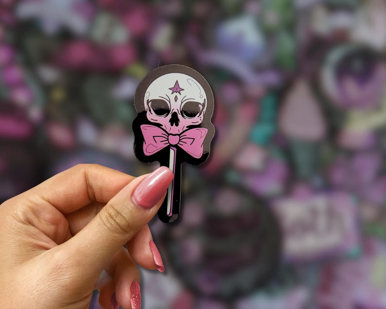 May include: A sticker featuring a skull-shaped lollipop with a pink bow tie. The skull is white with black eye sockets and a star on its forehead. The lollipop stick is black, and the background is a blur of purple and pink.