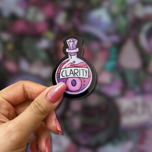 May include: A sticker featuring a potion bottle design with the word "CLARITY" in a black border. The bottle contains a pink liquid and a purple eye-shaped object. The bottle has a purple gem-like stopper.