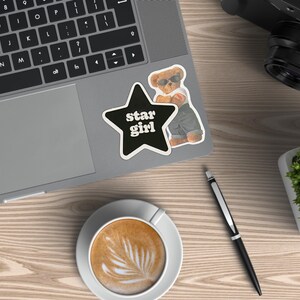 May include: A laptop with a black keyboard and a sticker that says "star girl" next to a cartoon bear wearing sunglasses. A cup of coffee with latte art, a pen, a camera, and a small potted plant are also on the wooden surface.