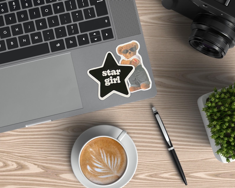 May include: A laptop with a black keyboard and a sticker that says "star girl" next to a cartoon bear wearing sunglasses. A cup of coffee with latte art, a pen, a camera, and a small potted plant are also on the wooden surface.