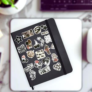 May include: A black notebook covered in various stickers, including stars, cherries, and hearts. The stickers feature text like "Stay Wild Moon Child" and "Rock N Roll". The notebook is on a white surface, with a computer mouse and keyboard in the background.