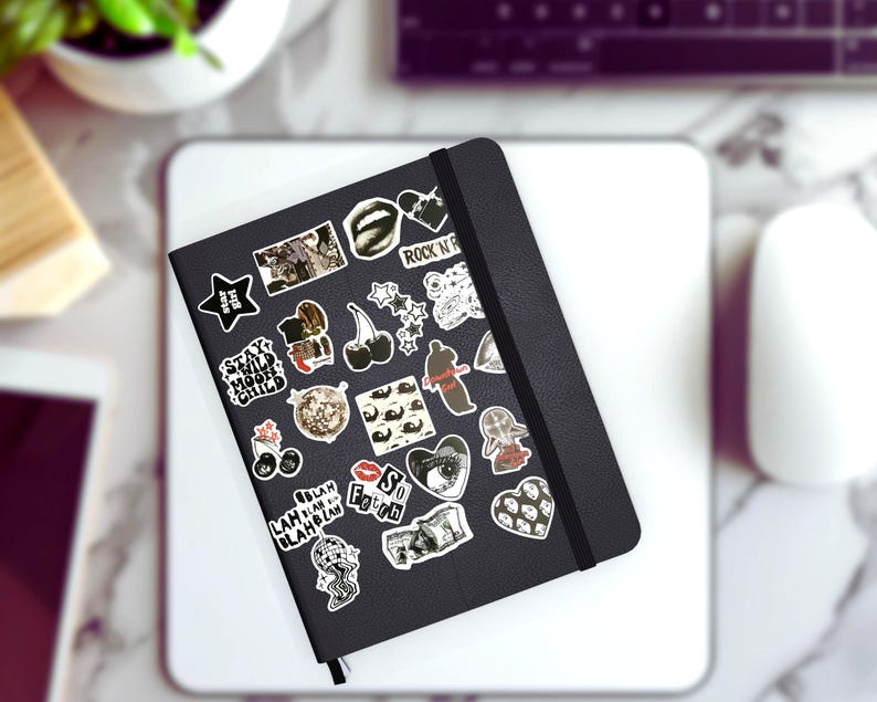 May include: A black notebook covered in various stickers, including stars, cherries, and hearts. The stickers feature text like "Stay Wild Moon Child" and "Rock N Roll". The notebook is on a white surface, with a computer mouse and keyboard in the background.