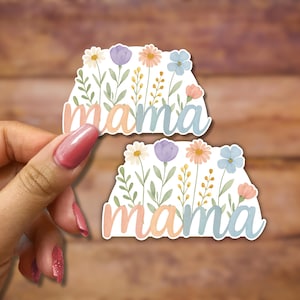 May include: Two white stickers with the word "mama" in colorful letters surrounded by watercolor flowers. The flowers are in shades of pink, blue, yellow, and green.