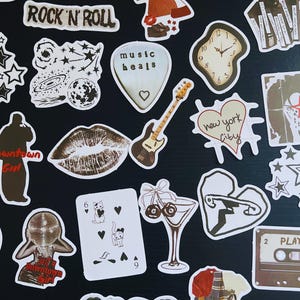 May include: A collection of assorted stickers with a rock and roll theme. Designs include a guitar, lips, a clock, stars, and the words "Rock 'N' Roll", "music heals", "Downtown Girl", and "New York City".
