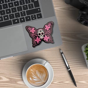 May include: A black butterfly sticker with a skull design and pink floral accents rests on a laptop. The sticker is next to a cup of coffee, a pen, a camera, and a small plant on a wooden surface.