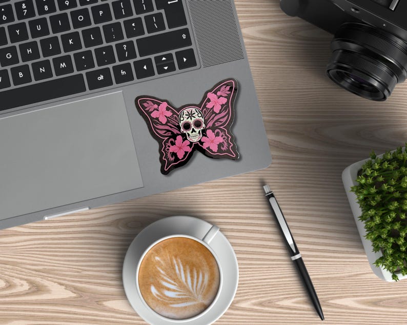 May include: A black butterfly sticker with a skull design and pink floral accents rests on a laptop. The sticker is next to a cup of coffee, a pen, a camera, and a small plant on a wooden surface.