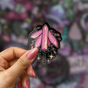 May include: A pink crystal sticker with a black outline. The crystal is the focal point, with a sword-like base and leafy accents. The sticker is held in a hand with pink nail polish, set against a blurred background of other stickers.