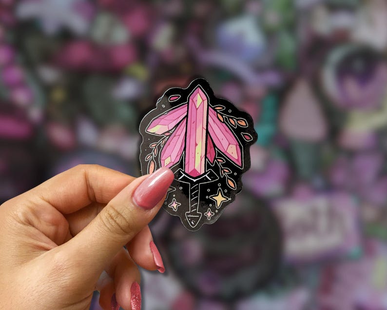May include: A pink crystal sticker with a black outline. The crystal is the focal point, with a sword-like base and leafy accents. The sticker is held in a hand with pink nail polish, set against a blurred background of other stickers.