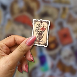 May include: A rectangular sticker featuring a vintage illustration of a skull in a vase with flowers. The sticker has a decorative border and is held against a blurred background of other stickers. The color palette includes beige, red, and brown.