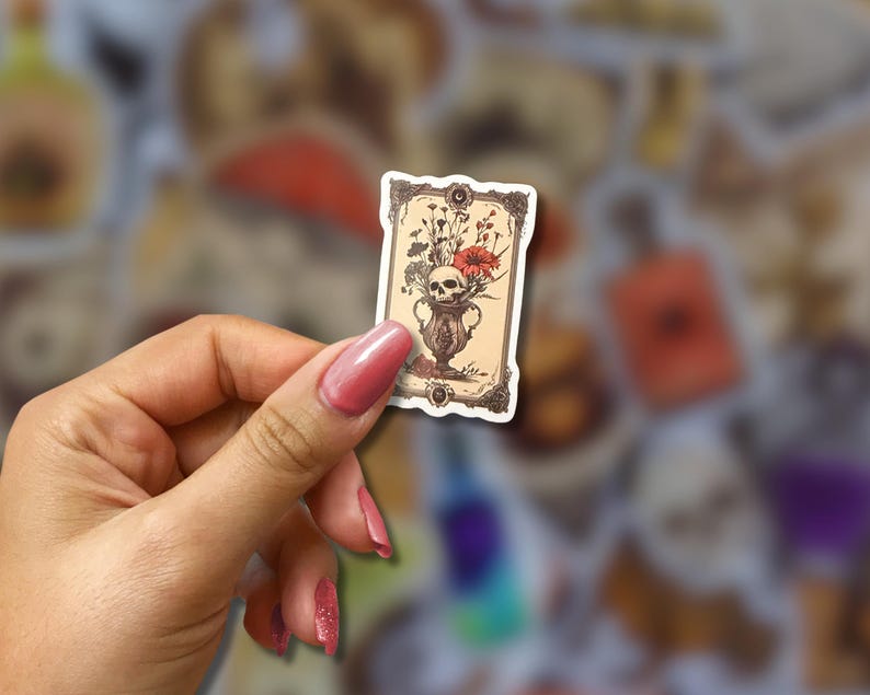 May include: A rectangular sticker featuring a vintage illustration of a skull in a vase with flowers. The sticker has a decorative border and is held against a blurred background of other stickers. The color palette includes beige, red, and brown.
