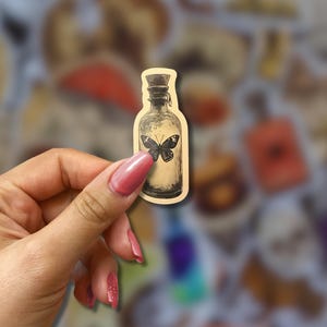 May include: A sticker featuring a vintage-style bottle with a butterfly illustration. The bottle is beige with a dark cap and a black butterfly in the center. The sticker has a slightly blurred background with other similar stickers.