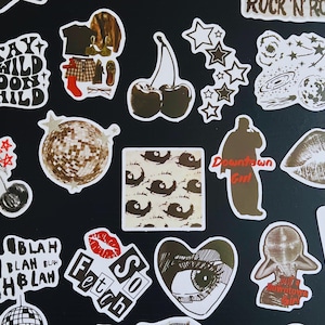 May include: A collection of various stickers with white borders on a black background. Designs include cherries, stars, a disco ball, lips, and text such as "Stay Wild Moon Child" and "So Fetch". The stickers are in shades of brown, white, and red.