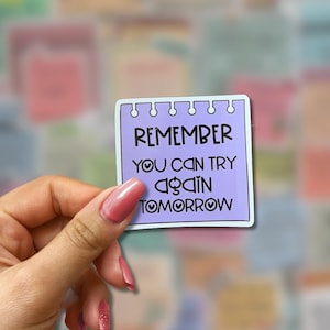 May include: A square sticker with a light purple background and white border. The sticker has the words "REMEMBER YOU CAN TRY AGAIN TOMORROW" in black. The sticker is held by a hand with pink nail polish.