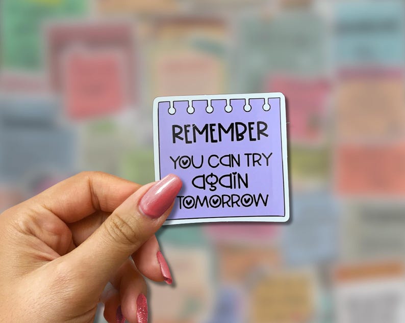 May include: A square sticker with a light purple background and white border. The sticker has the words "REMEMBER YOU CAN TRY AGAIN TOMORROW" in black. The sticker is held by a hand with pink nail polish.