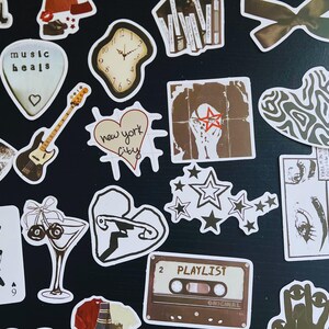 May include: A collection of assorted stickers with various designs. Includes a guitar, a heart with "new york city" text, a cassette tape labeled "PLAYLIST", a clock, and a lips design. The stickers are in shades of brown, white, and black.