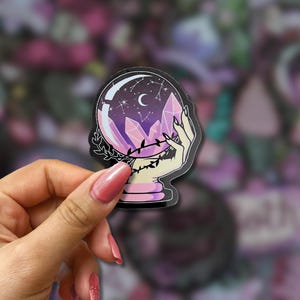May include: A sticker featuring a crystal ball with a crescent moon and stars inside. The crystal ball is held by a hand with long nails, and surrounded by purple crystals. The sticker has a black outline and a pink and purple color scheme.