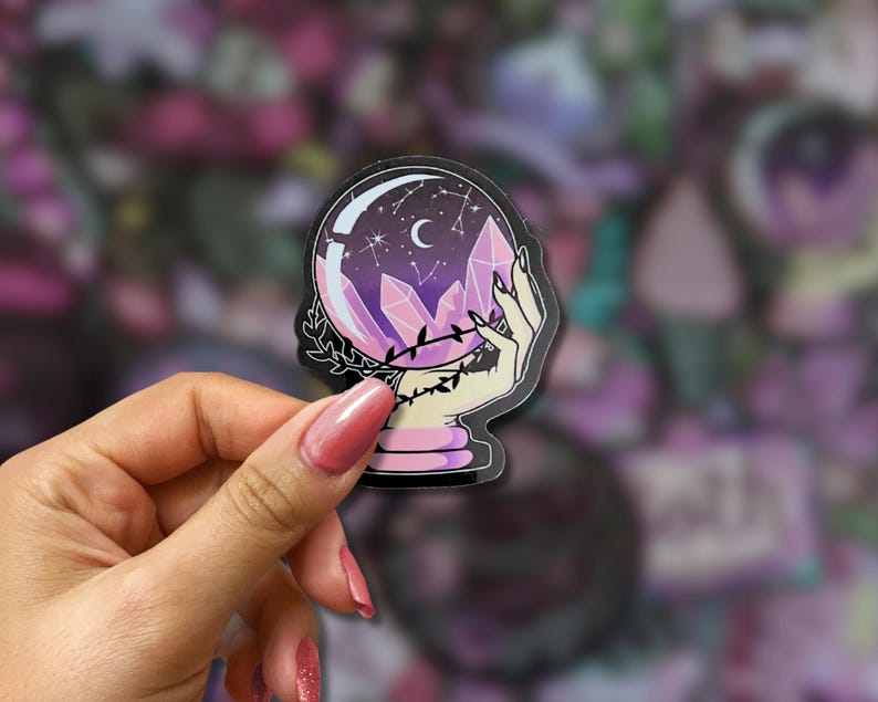 May include: A sticker featuring a crystal ball with a crescent moon and stars inside. The crystal ball is held by a hand with long nails, and surrounded by purple crystals. The sticker has a black outline and a pink and purple color scheme.