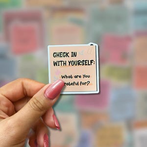 May include: A square sticker with the text "CHECK IN WITH YOURSELF: What are You grateful for?" in black font. The sticker has a light peach background with a light blue border and a small floral design. The sticker is held by a hand.