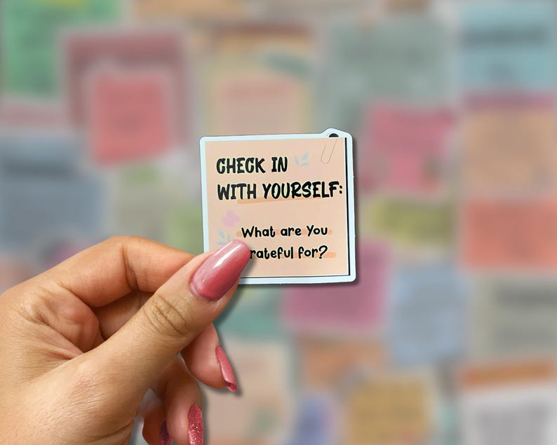 May include: A square sticker with the text "CHECK IN WITH YOURSELF: What are You grateful for?" in black font. The sticker has a light peach background with a light blue border and a small floral design. The sticker is held by a hand.