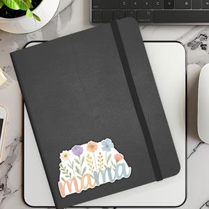 May include: A black leather journal with a floral sticker that says "mama". The sticker features pink, blue, and yellow flowers with green leaves.