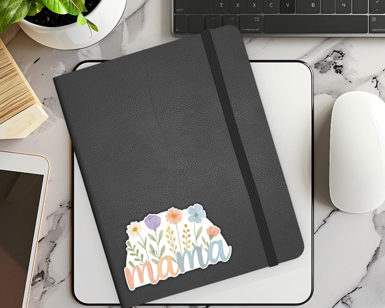 May include: A black leather journal with a floral sticker that says "mama". The sticker features pink, blue, and yellow flowers with green leaves.