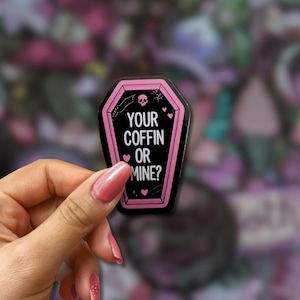May include: A black coffin-shaped sticker with a pink border and the text "YOUR COFFIN OR MINE?". The sticker features a skull, hearts, and stars. The sticker is held in a hand.
