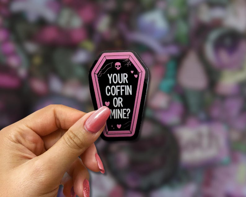 May include: A black coffin-shaped sticker with a pink border and the text "YOUR COFFIN OR MINE?". The sticker features a skull, hearts, and stars. The sticker is held in a hand.