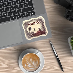 May include: A sticker on a laptop featuring a vintage illustration of a woman holding a glass of wine, with a cat. The word "WOMAN" is at the top of the sticker. The sticker is on a laptop on a wooden desk, with a cup of coffee and a pen.