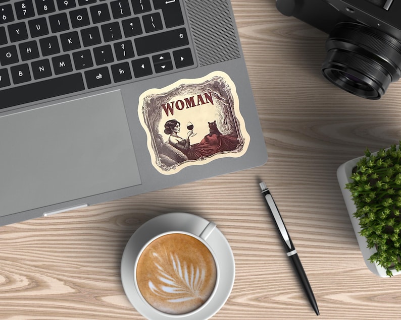 May include: A sticker on a laptop featuring a vintage illustration of a woman holding a glass of wine, with a cat. The word "WOMAN" is at the top of the sticker. The sticker is on a laptop on a wooden desk, with a cup of coffee and a pen.