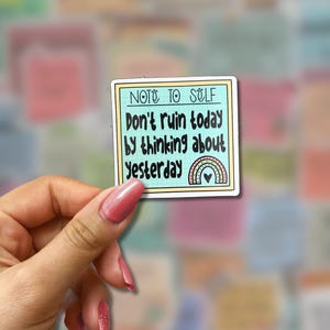 May include: A square sticker with a light blue border and the text "NOTE TO SELF" at the top. The sticker reads "Don't ruin today by thinking about yesterday" with a rainbow and heart design. The sticker is held by a hand.