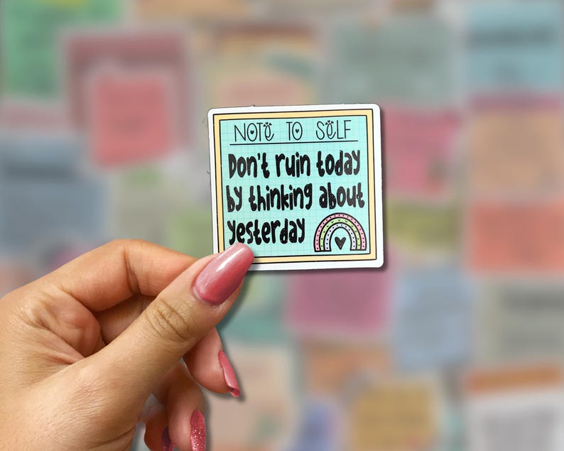 May include: A square sticker with a light blue border and the text "NOTE TO SELF" at the top. The sticker reads "Don't ruin today by thinking about yesterday" with a rainbow and heart design. The sticker is held by a hand.