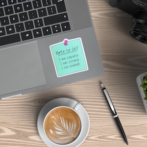 May include: A laptop with a keyboard and a sticky note that reads "Note to Self: I am capable, I am strong, I am enough." A cup of coffee, a pen, a camera, and a small plant are also on the wooden surface.