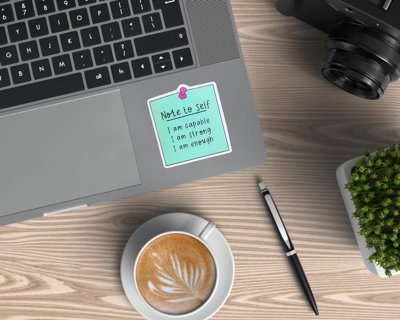 May include: A laptop with a keyboard and a sticky note that reads "Note to Self: I am capable, I am strong, I am enough." A cup of coffee, a pen, a camera, and a small plant are also on the wooden surface.
